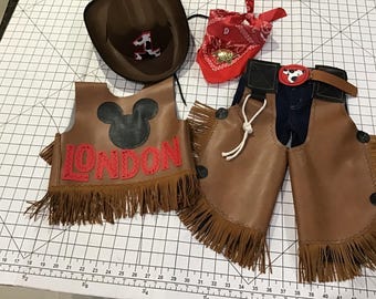 Puede incluir: Un conjunto de disfraz de vaquero marrón que incluye sombrero, chaleco y polainas. El chaleco tiene una cabeza de Mickey Mouse negra y la palabra "LONDON" en rojo. Las polainas tienen flecos y un cinturón con un gráfico de vaca. También se incluye un pañuelo rojo.