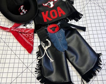 May include: A black cowboy hat with a red and white emblem, a red bandana, and a black vest with the letters "KOA" in red. The outfit includes denim chaps with black fringe and a belt with a red buckle.