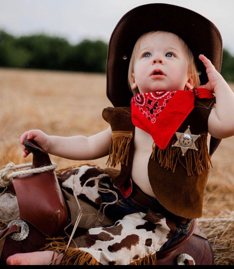 May include: A baby wearing a brown cowboy hat, a red bandana, a brown vest with fringe, and a pair of jeans with a cow print pattern. The baby is sitting on a brown saddle.