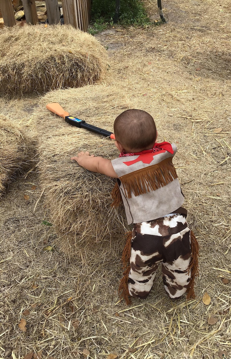 May include: A young child dressed in a brown cowboy vest with fringe, a red bandana, and brown and white cow print pants. The child is holding a toy gun and standing in a pile of hay.