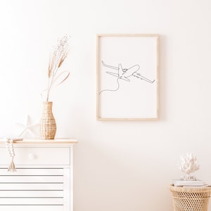 May include: A framed black and white line drawing of an airplane in flight. The drawing is on a white background and is framed in a light wood frame. The frame is hanging on a white wall.