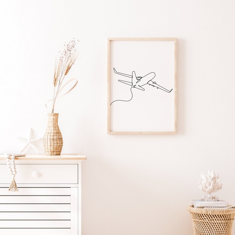 May include: A framed black and white line drawing of an airplane in flight. The drawing is on a white background and is framed in a light wood frame. The frame is hanging on a white wall.