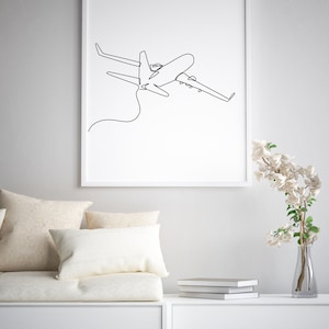 May include: A white framed print of a black line drawing of an airplane in flight. The print is hanging on a white wall above a white couch with beige pillows. A vase with white flowers is on a white shelf to the right of the couch.