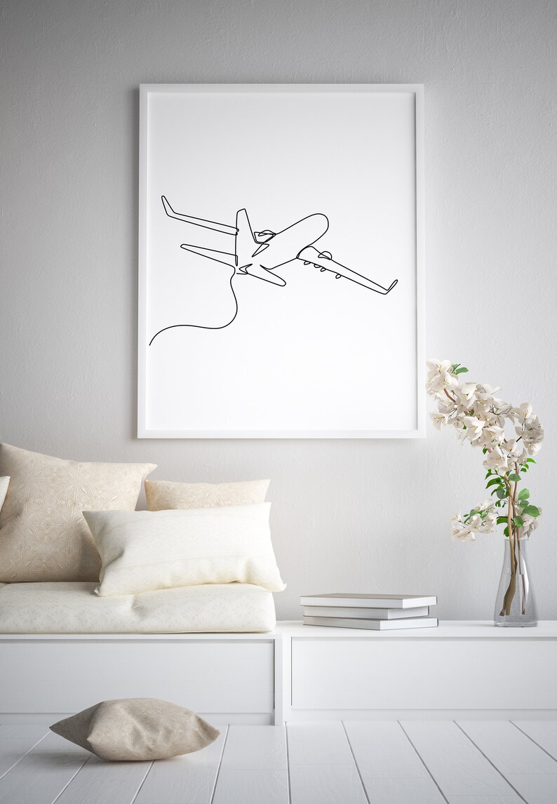 May include: A white framed print of a black line drawing of an airplane in flight. The print is hanging on a white wall above a white couch with beige pillows. A vase with white flowers is on a white shelf to the right of the couch.