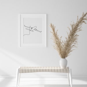 May include: A white bench with a woven seat and white legs sits in front of a white wall. A white frame with a black line drawing of an airplane hangs on the wall. A vase with dried pampas grass sits on the bench.