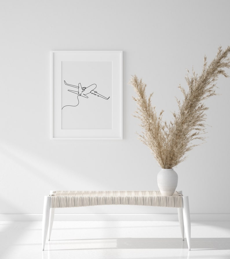 May include: A white bench with a woven seat and white legs sits in front of a white wall. A white frame with a black line drawing of an airplane hangs on the wall. A vase with dried pampas grass sits on the bench.