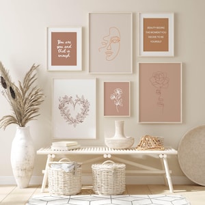May include: A white bench with two white wicker baskets underneath. There are five framed prints on the wall above the bench. The prints feature line drawings of flowers, faces, and inspirational quotes. "You are you and that is enough" and "Beauty begins the moment you decide to be yourself" are the quotes on the prints.