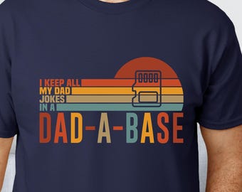 May include: Navy blue t-shirt with a retro design. The graphic features a sunset with orange, yellow, and green stripes, a memory card icon, and the text "I KEEP ALL MY DAD JOKES IN A DAD-A-BASE."