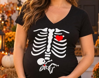May include: A black V-neck t-shirt featuring a white graphic print of a human rib cage with a red heart on the left side and a small skeleton figure positioned near the bottom, representing a baby skeleton with a tiny red heart.