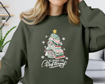Puede incluir: Una sudadera verde con un árbol de Navidad hecho de libros apilados. El árbol está decorado con luces y una estrella en la cima. El texto "All Booked for Christmas" está impreso debajo del árbol.