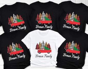 Puede incluir: Una colección de camisetas negras y blancas y una camiseta blanca. Cada camiseta presenta una camioneta roja que transporta un árbol de Navidad verde, con un bosque de árboles en el fondo. El texto "Brown Family" está impreso debajo.