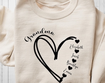 Könnte beinhalten: Ein cremefarbener Pullover mit dem Wort "Grandma" in Schwarz gedruckt über einem handgezeichneten Herzen. Im Herzen sind die Namen "Elizabeth", "Jacob", "Brooklyn" und "Emma" in Schwarz gedruckt mit einem kleinen schwarzen Herzen neben jedem Namen.