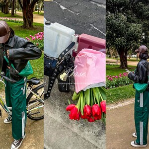 May include: A person wearing a black leather jacket, green Adidas track pants with white stripes, and black Converse sneakers. They are holding a green purse and a brown cap. A bicycle is in the foreground, and a bouquet of red tulips is visible.