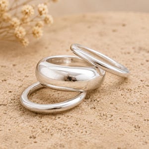 Puede incluir: Un conjunto de tres anillos plateados pulidos apilados. La colección incluye una banda ancha y redondeada colocada en el centro, flanqueada por dos bandas circulares más delgadas y lisas. La superficie del metal es reflectante y uniforme.