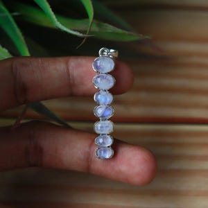 Puede incluir: Un colgante de plata con siete piedras preciosas de ópalo lunar ovaladas. Las piedras lunares tienen una base blanca lechosa con un brillo azul iridiscente. El colgante está sostenido por una mano sobre un fondo de madera.