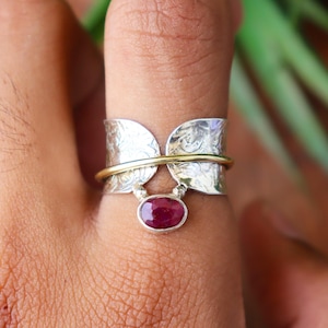 May include: A silver ring with a gold band and a dangling oval-shaped, deep red gemstone. The ring has a unique design with etched patterns on the silver portions, creating an elegant and eye-catching piece of jewelry.