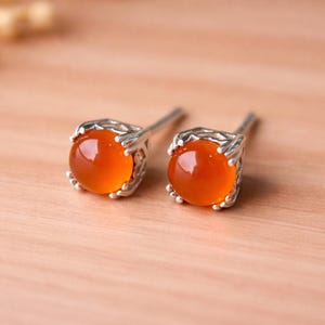 May include: A pair of stud earrings featuring round, orange-colored gemstones set in ornate silver-toned metal. The earrings have a post backing and are displayed on a light brown wooden surface. These earrings are a jewelry accessory.