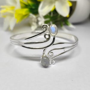 以下が含まれることがあります： 2つの楕円形のムーンストーンアクセントが付いたシルバーカラーのブレスレット。ブレスレットは、渦巻きの装飾要素を備えた繊細なオープンデザインです。ムーンストーンは微妙な虹色の輝きがあり、柔らかくぼやけた背景に設定されています。