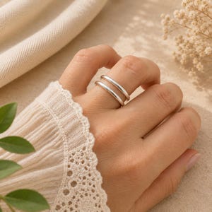 May include: A double band ring made of polished silver. The ring features two smooth, parallel bands that are connected, creating a layered appearance on the finger. The metal has a bright, reflective finish that catches the light.