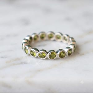 May include: A silver eternity band ring featuring a continuous row of round, faceted olive-green gemstones set in individual circular metal bezels. The ring has a polished metallic finish and a uniform design that encircles the entire band.