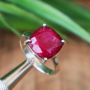 May include: A silver ring featuring a large, cushion-cut ruby gemstone. The ruby is a vibrant red color and is set in a four-prong setting. The ring's band is a simple, polished silver. The background is blurred green.