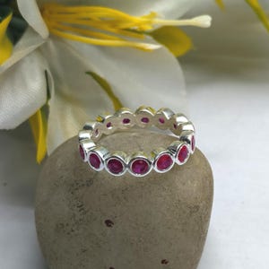 May include: A silver eternity band ring featuring a continuous row of round, vibrant pink stones set in individual circular bezels. The ring is positioned upright, showcasing the uniform arrangement of the stones and the polished metal finish.