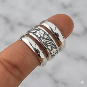 May include: A set of three silver rings stacked together. The outer two rings have a smooth, polished, reflective surface. The centre ring features a detailed, embossed floral pattern with small flowers and leaves set against a dark background.