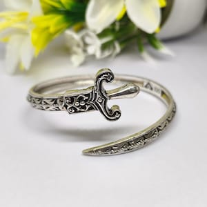 May include: A silver-colored bracelet with an ornate sword design. The sword features intricate detailing and is the focal point of the open-ended cuff. The bracelet has a decorative pattern along its band.