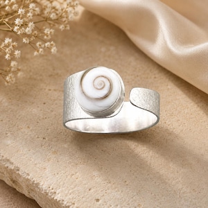 Könnte beinhalten: Ein silberfarbener Metallring mit offenem Banddesign und gebürsteter Oberfläche. Das Herzstück ist eine kreisförmige Einlage aus weißer Muschel mit einem natürlichen braunen Spiralmuster, die vorne auf dem verstellbaren Metallband befestigt ist.