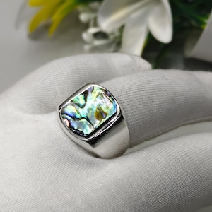 May include: A silver-toned ring featuring a rectangular inlay of iridescent abalone shell. The shell displays a vibrant mix of green, blue, and purple hues with a polished, reflective surface. The metal band is smooth, wide, and has a high-shine finish.