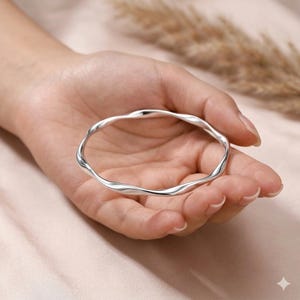 May include: A polished silver-toned bangle bracelet featuring a continuous, smooth, undulating wave pattern. The circular jewellery piece has a reflective metallic surface with gentle curves that create a fluid, rippled appearance around the entire band.