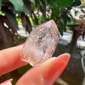 Könnte beinhalten: Ein klarer Quarz-Kristall mit einer Spitze. Der Kristall wird vor einem Hintergrund aus grünem Laub und einem Fenster gehalten. Der Kristall hat einen leicht bräunlichen Farbton und erscheint durchscheinend.