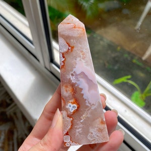 May include: A pink and orange agate crystal point with white and brown inclusions. The crystal is shaped like a pyramid and has a smooth, polished surface.