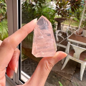 Pode incluir: Uma ponta de cristal de quartzo rosa transparente e rosa. O cristal é segurado em frente a uma janela com folhagem verde exuberante ao fundo. O cristal tem um topo facetado e pontiagudo e uma base retangular.