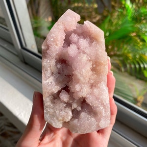 May include: A light pink crystal cluster with a rough, textured surface. The crystal has a rectangular shape with a pointed top and a hollowed-out center. The crystal is held in a hand.