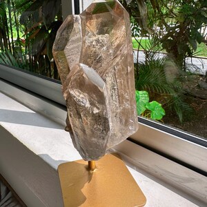 May include: A large, clear quartz crystal cluster with a golden brown hue, displayed on a gold metal stand. The crystal has a natural, rough texture and is illuminated by sunlight.