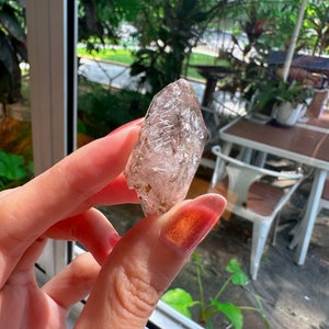 Könnte beinhalten: Ein klarer Quarzkristall mit einer spitzen Spitze und einer rauen, natürlichen Textur. Der Kristall wird in einer Hand mit rosafarbenem Nagellack gehalten.