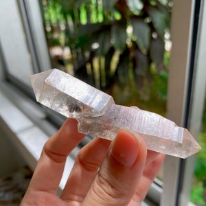 May include: A clear quartz crystal with a double-terminated point, also known as a double-ended crystal. The crystal is held in a hand, showcasing its translucent and slightly cloudy appearance.