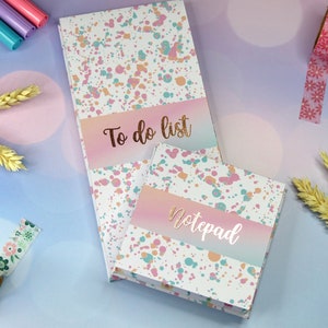 May include: Two notebooks with a white background and colorful splatter paint design. The larger notebook has the text "To do list" in gold lettering. The smaller notebook has the text "Notepad" in gold lettering.