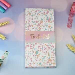 Könnte beinhalten: Ein weißes Notizbuch mit der Aufschrift "To do list" mit einem pinkfarbenen, blauen und orangen Spritzmuster. Der Einband hat einen goldenen Schriftzug "To do list".