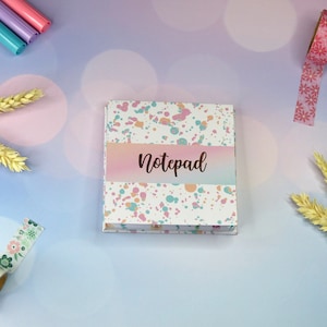 Puede incluir: Un bloc de notas blanco con un patrón de salpicaduras rosa, azul y naranja. El bloc de notas tiene una banda degradada rosa, naranja y azul en la parte delantera con la palabra "Notepad" escrita en texto marrón.