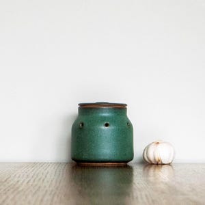 Op de afbeelding: Een groene keramische knoflookhouder met een deksel en drie kleine gaatjes, naast een knoflookbol. De houder heeft een donkerbruine rand en basis. De items staan op een houten oppervlak tegen een witte achtergrond.