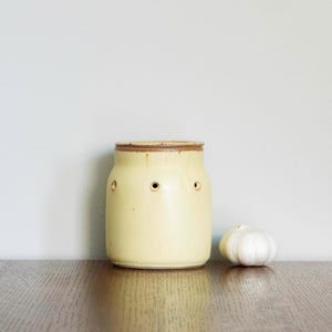 Może przedstawiać: Jasnożółty ceramiczny pojemnik na czosnek z pokrywką i trzema małymi otworami. Cała główka czosnku leży obok pojemnika na drewnianej powierzchni. Pojemnik ma rustykalny, ręcznie robiony wygląd.
