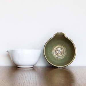 May include: Two ceramic bowls on a wooden surface. A white bowl with a spout is on the left. A green bowl with a grater surface is on the right. The bowls are for kitchen use.