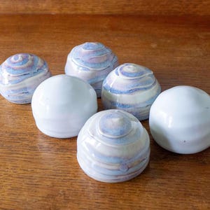 May include: A collection of six small, rounded ceramic objects. Three of the objects feature a smooth, solid white glaze, while the other three display a swirling, marbled pattern of light blue, lavender, and white tones across their surfaces.