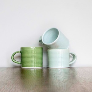 Puede incluir: Tres tazas de cerámica apiladas sobre una superficie de madera. La taza superior es de color azul claro, la taza del medio es de color azul claro y la taza inferior es de color verde.