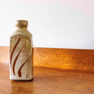 Puede incluir: Una botella de cerámica beige con forma rectangular y cuello estrecho. La botella tiene vetas y motas marrones. Se encuentra sobre una superficie de madera.