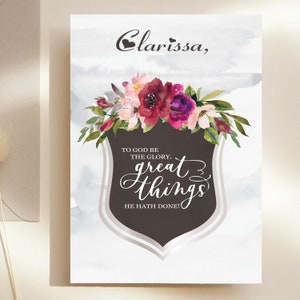 May include: A watercolour floral design with a brown shield featuring the text "To God be the glory, great things He hath done!" and the name "Clarissa" at the top.