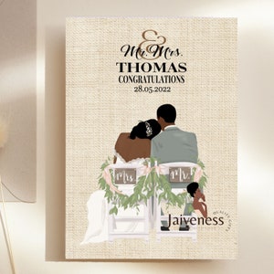 Peut inclure: Une carte de mariage avec un fond en toile de jute repr&eacute;sentant un couple noir assis sur des chaises avec de la verdure et des pancartes indiquant "Mr." et "Mrs.". Le texte sur la carte est "Mr. & Mrs. Thomas F&eacute;licitations 28.05.2022".