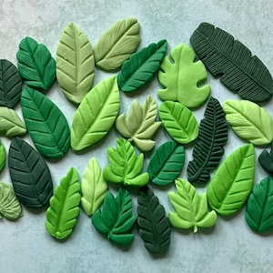 May include: A collection of 20 green leaf shapes made of polymer clay. The leaves are in various sizes and shapes, with some having a more realistic texture and others being more stylized.